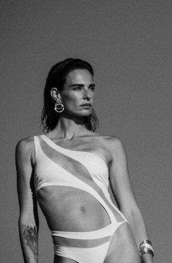 Pharos One-Piece 30 Back pose on rocky shore displaying Pharos swimsuits intricate ring strapped open design in white