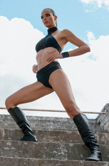 Athena SwimWear 23 ATHENA CROP TOP in black intricate lace embroidery highlights toned abs as model reclines on ancient stone steps with raised arm pose