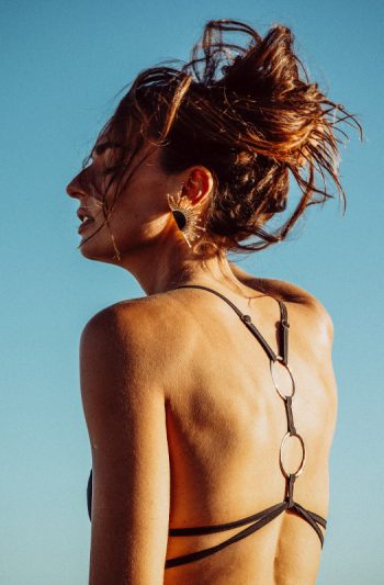 Rhea Bikini 25 RHEA black bikini with gold accents and adjustable ties sensual pose with arms raised against ocean backdrop
